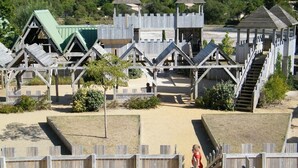 Eine hölzerne Spielanlage mit mehreren Rutschen und Kletterbereichen, umgeben von Sand und Grünpflanzen.