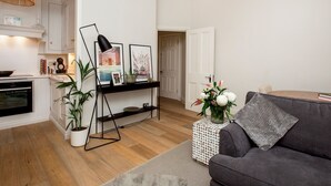 A modern living room with a grey sofa, a coffee table, and a black floor lamp. There is a white console table with framed pictures and a potted plant. The room has wooden flooring and white walls.