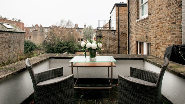 A patio area with a table and two wicker chairs, a vase with white flowers, and brick buildings in the background.
