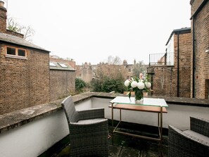 A rooftop terrace with wicker furniture, a glass table, and a vase of flowers.