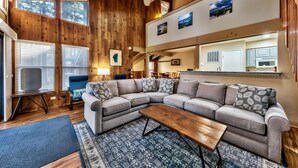 Ein gemütliches Wohnzimmer mit einem Ecksofa, einem Couchtisch und einem Fernseher. Holzverkleidungen, ein Fenster und eine Küche sind im Hintergrund zu sehen.