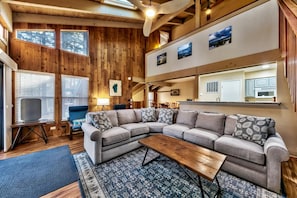 A cozy living room with a sectional sofa, a coffee table, and a TV. There are wooden walls, a window, and a kitchen visible in the background.