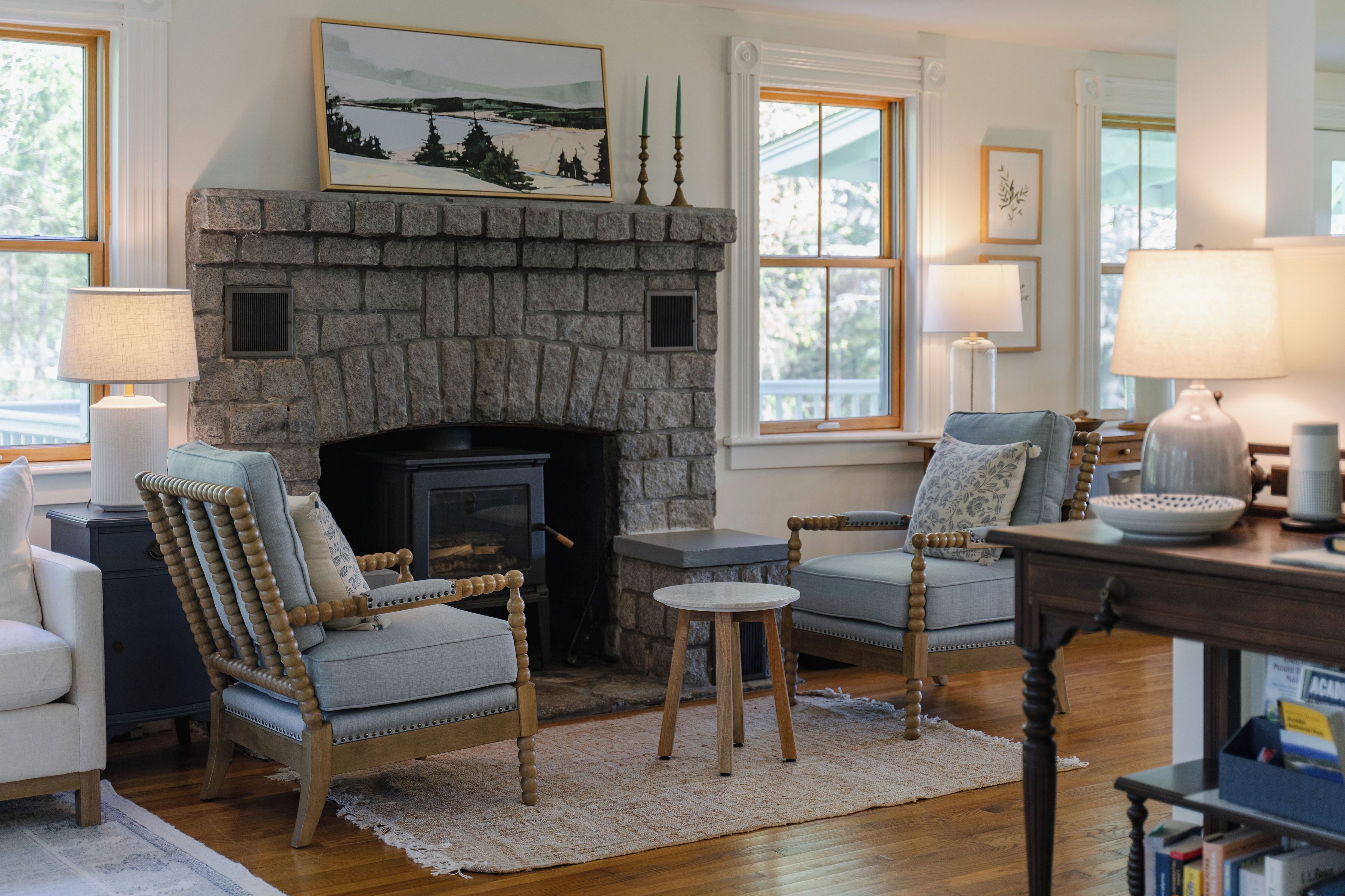 A cozy living room with a stone fireplace, two upholstered chairs, a small round table, and a lamp on a wooden table.