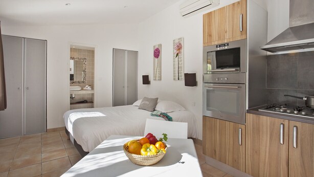 Une chambre moderne avec un grand lit, une table de chevet sur laquelle se trouve un panier de fruits, et une kitchenette équipée d’une cuisinière et d’un four.