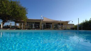 Une piscine avec une eau d’un bleu limpide, un bâtiment doté d’un toit incurvé et un arbre sur la gauche.