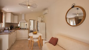 A compact kitchen with white cabinets, a white countertop, a dining table with chairs, a hanging light fixture, and a round mirror on the wall. - maison kiwi avec piscine chauffée  terrasse  jardin et bbq à Calvi (Calvi)