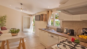A modern kitchen with a gas stove, a sink, and a window with curtains. There is a dining area with a table and chairs, and a plant on the table. - maison kiwi avec piscine chauffée  terrasse  jardin et bbq à Calvi (Calvi)