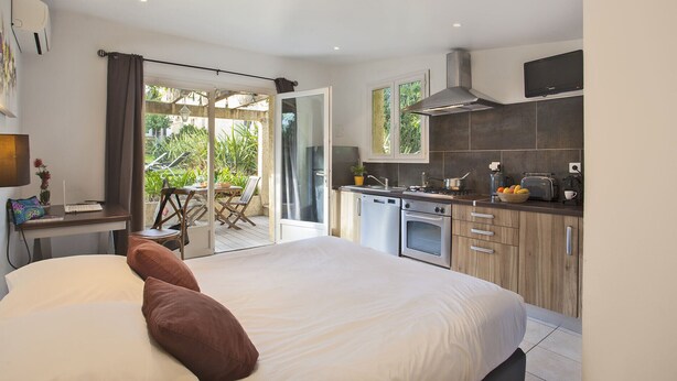 A modern bedroom with a large bed, a small kitchenette, and a sliding glass door leading to a balcony. - minivilla lilas indépendante à Calvi avec jardin et piscine, jardin et bbq (Calvi)