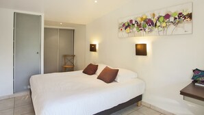 A bedroom with a bed, two wall-mounted lights, a wooden chair, and a colorful abstract painting. - minivilla lilas indépendante à Calvi avec jardin et piscine, jardin et bbq (Calvi)