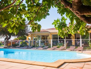 Un área junto a la piscina con sillones de descanso, una piscina azul cristalina y un edificio con grandes ventanales y tejado de tejas.