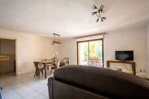 A modern living room with a sofa, a dining area with chairs and a table, a TV on a stand, and a ceiling light fixture.