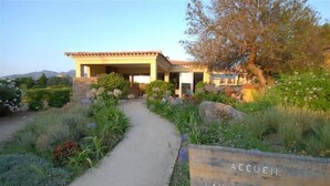 Ein Gebäude mit einer überdachten Terrasse und einem Weg, der dorthin führt, umgeben von einem Garten mit verschiedenen Pflanzen und einem Schild mit der Aufschrift 'ACCEIL LAURIERS ROSES'.