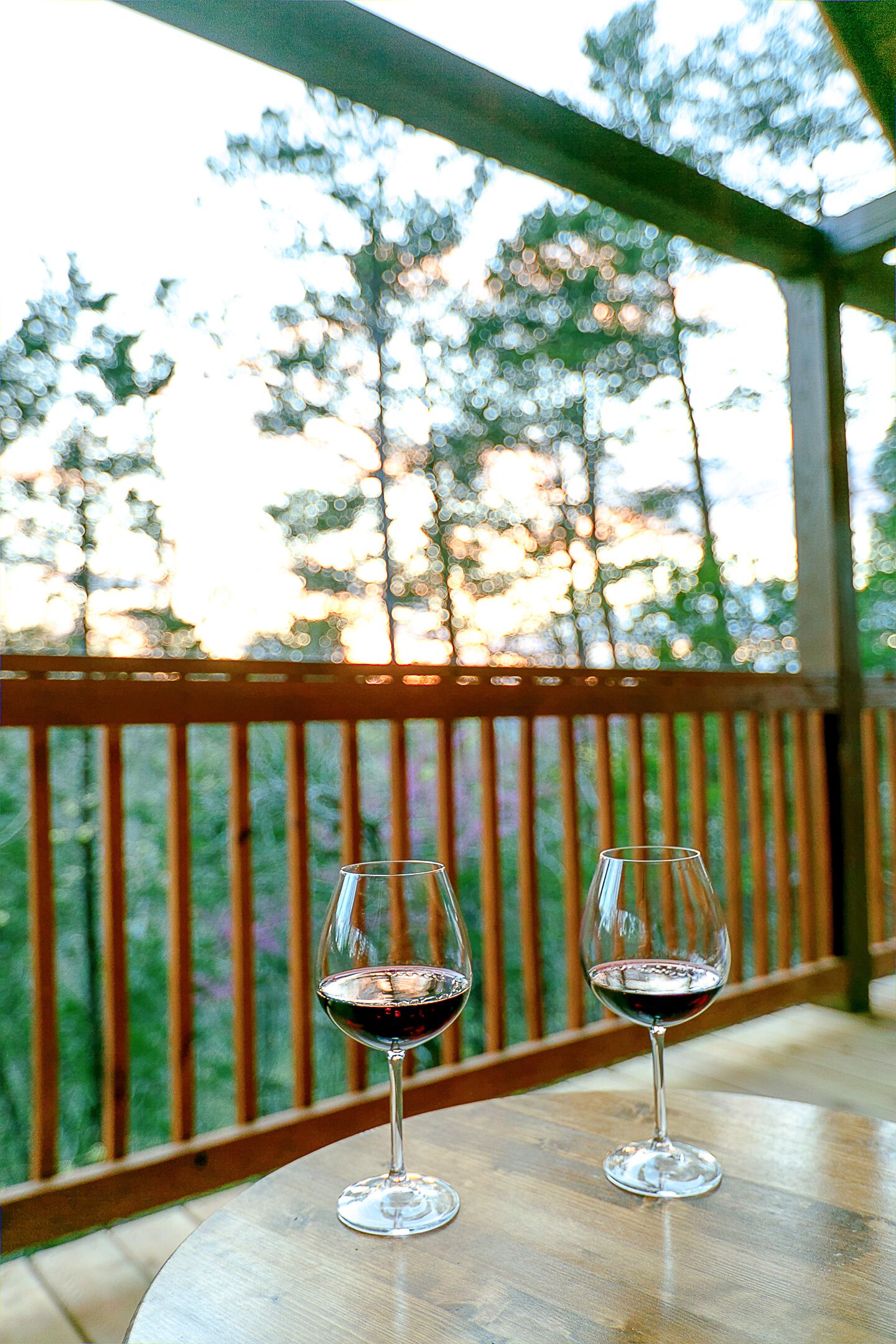 Deux verres à vin sont posés sur une table en bois, et on aperçoit des arbres à travers une rambarde en bois.