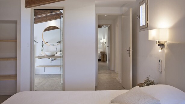 A modern hotel room with a large bed, a bathroom with a round mirror, and a hallway leading to another room.