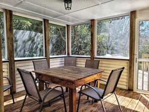 Een overdekt terras met een houten tafel en stoelen, grote ramen en uitzicht op bomen.