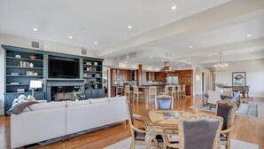 Uma sala de estar ampla com um sofá grande, uma mesa de jantar com cadeiras e uma área de cozinha ao fundo.