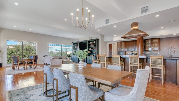 Uma sala de estar ampla, com uma grande mesa de jantar de madeira, cadeiras estofadas cinzentas e um lustre.