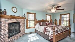 A bedroom with a brick fireplace, a wooden bed with patterned bedding, and two windows with views of greenery. - Get away from it all (Foristell)