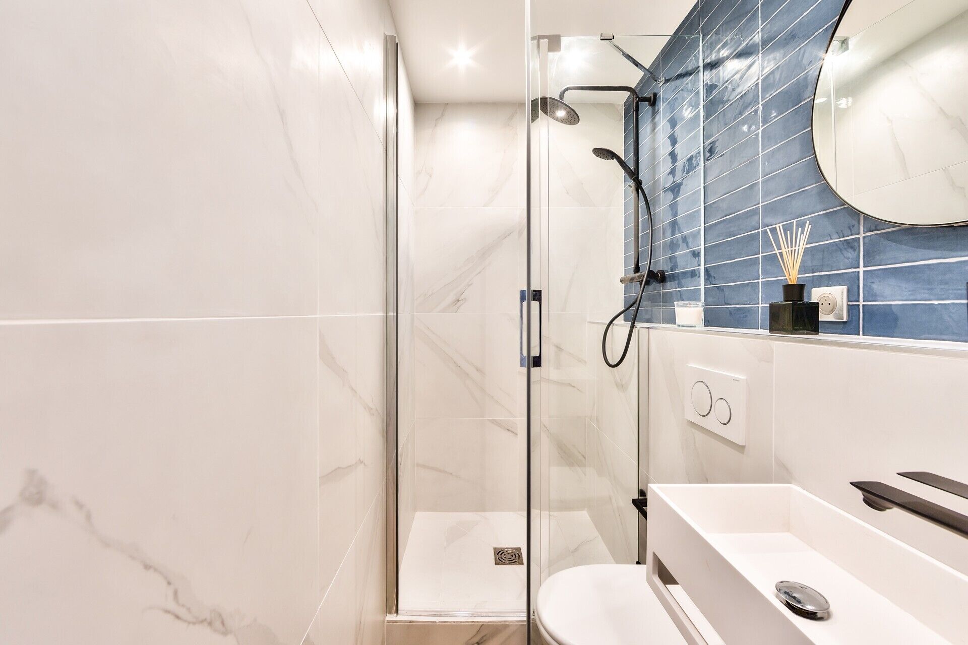 A modern bathroom with a glass-enclosed shower, white marble tiles, a blue-tiled wall, and a white sink with a black faucet.