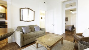 Ein Wohnzimmer mit Holztisch, einer Couch mit Kissen, einem Spiegel an der Wand und einem durchgehenden Türrahmen sichtbaren Küchenbereich.