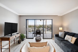 A modern living room with a flat-screen TV, a sofa, a coffee table, and a balcony with a view.