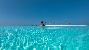 Mar azul e transparente com uma pessoa nadando, uma praia de areia e vários guarda-sóis.