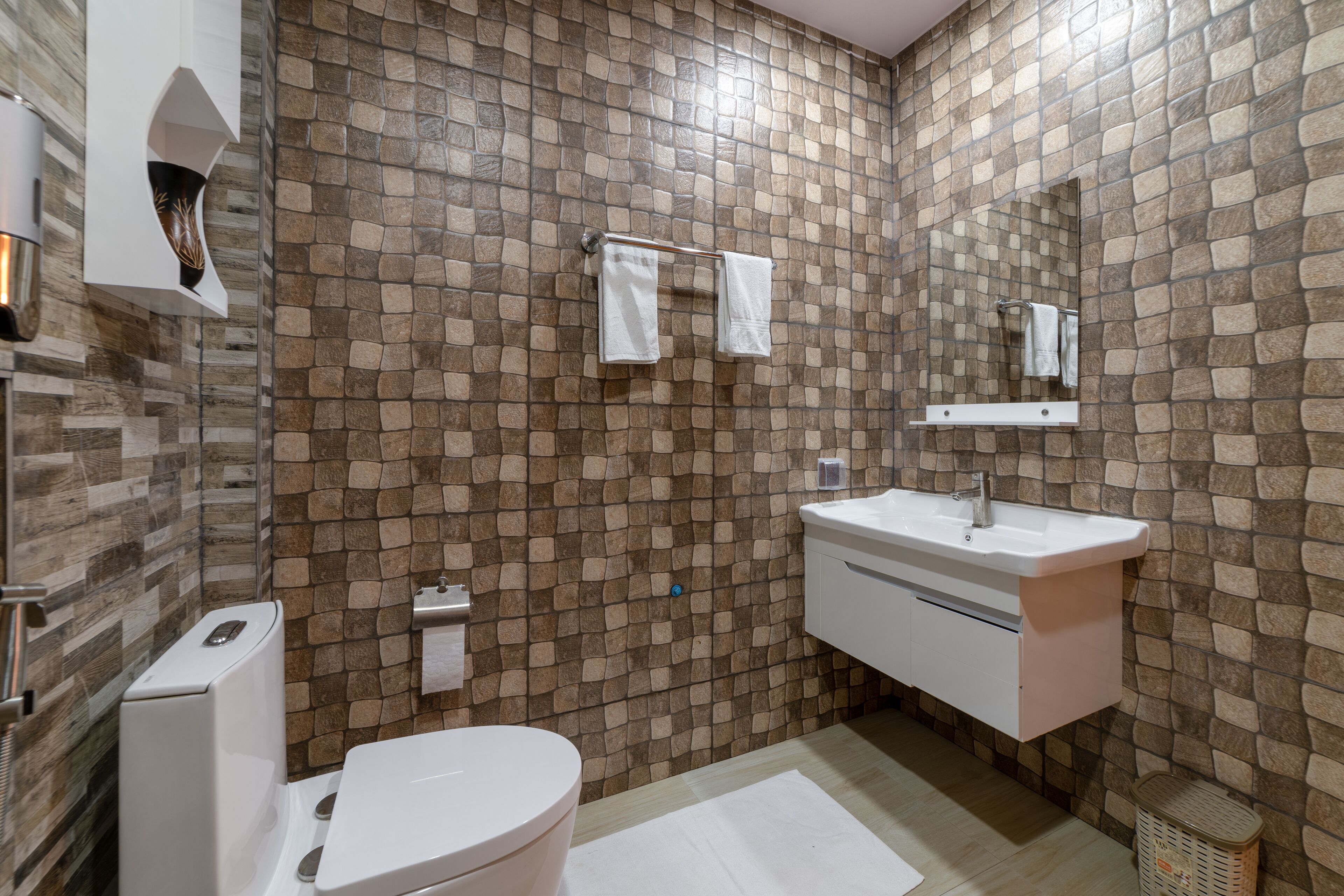 Ein Badezimmer mit einer Wand im Steinmuster, einer weißen Toilette, einem Waschbecken mit Spiegel und einem Handtuchhalter.