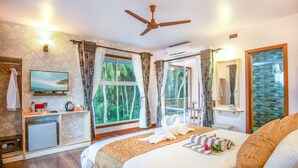 Quarto de hotel com cama, televisão, escrivaninha com computador, janela com cortinas e ventoinha no teto.