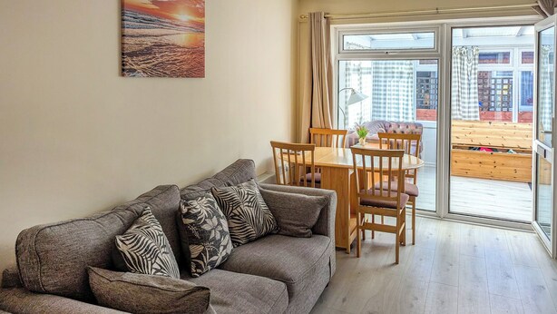 A living room with a sofa, a dining table, and chairs. There is a large window with curtains, and a painting on the wall. - 2 bedroom accommodation in Hunstanton (Hunstanton)