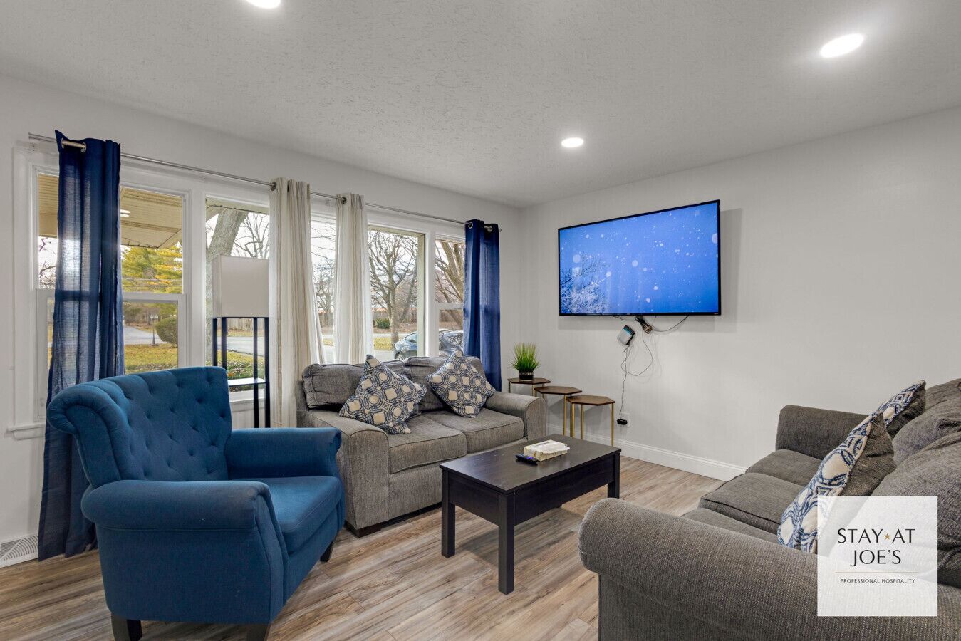 A modern living room with a blue armchair, grey sofa, and a coffee table. A flat-screen TV is mounted on the wall, and there is a sign for 'STAY AT JOE'S PROFESSIONAL HOSPITALITY'.