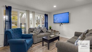 A modern living room with a blue armchair, grey sofa, and a coffee table. A flat-screen TV is mounted on the wall, and there is a sign for 'STAY AT JOE'S PROFESSIONAL HOSPITALITY'.
