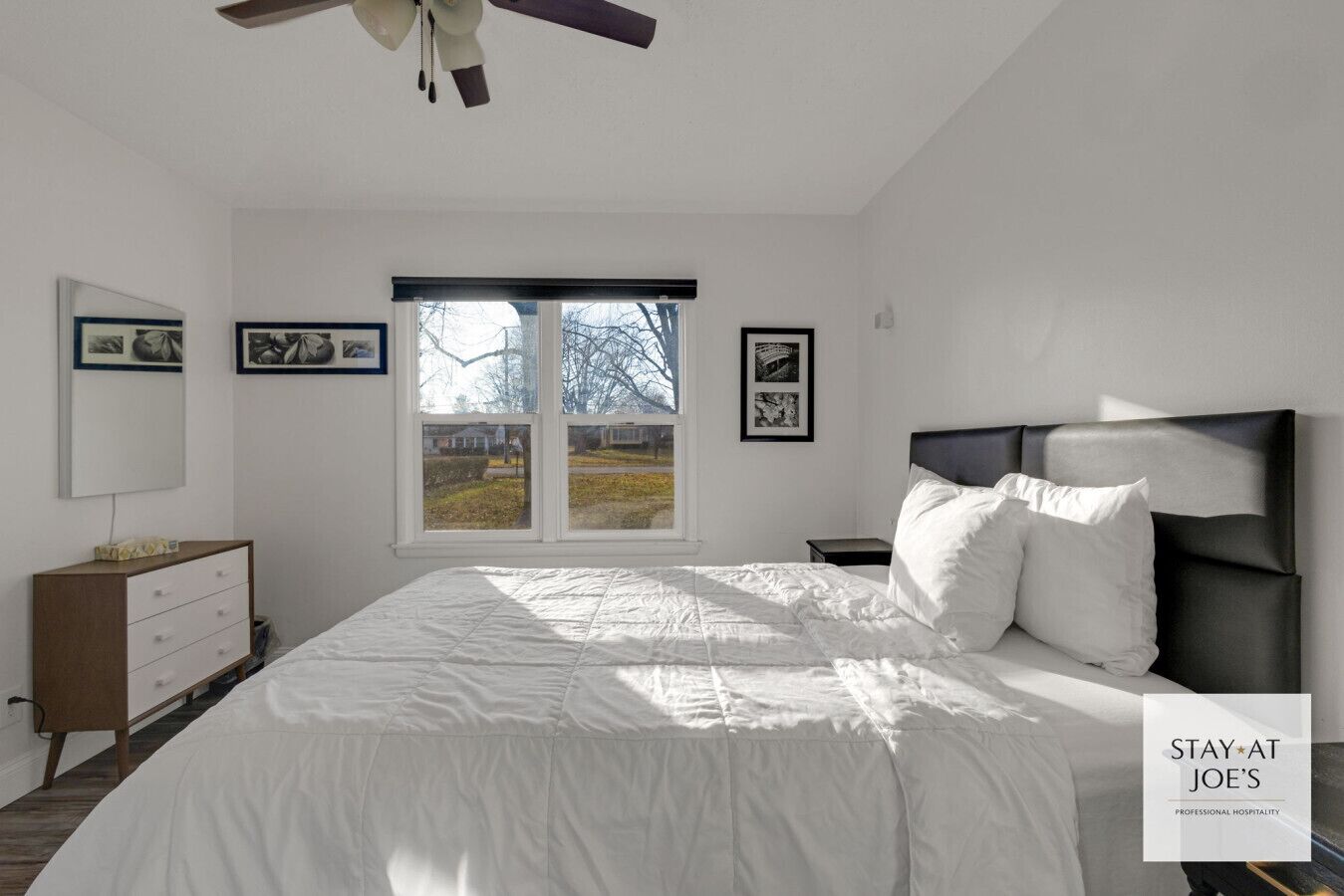 A bedroom with a large bed, a nightstand, a dresser, a ceiling fan, and a window with a view of trees.