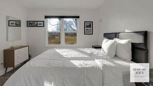 A bedroom with a large bed, a nightstand, a dresser, a ceiling fan, and a window with a view of trees.