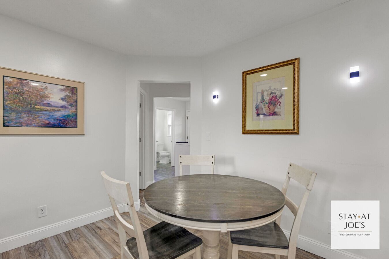 A dining area with a round table and four chairs, a painting on the wall, and a framed picture above the table.