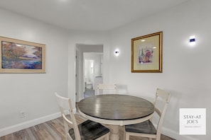 Uma área de refeições com uma mesa redonda e quatro cadeiras, um quadro na parede e uma moldura acima da mesa.