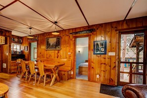 A wooden interior with a dining table, chairs, bookshelves, and a bed.