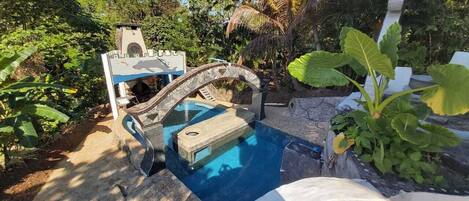 Una piscina con un puente de piedra, rodeada de abundante vegetación y una pequeña estructura de torre.