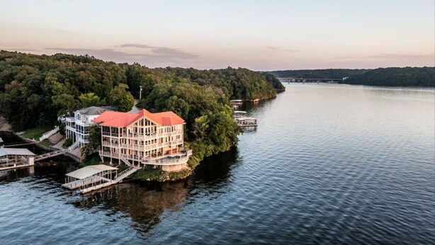 A large, multi-story building with a red roof, situated on a lakeside property with docks and a bridge in the background. - Allie's - Amazing 8,000 SF Home with --Private Pool--hot tub-- Sleeps 26+  (Sunrise Beach)