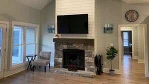 A living room with a stone fireplace, a ceiling fan, a flat-screen TV mounted above the fireplace, and a clock on the wall. - Beautiful waterfront home on a private cove in Lake Keowee w/ hot tub (Seneca)
