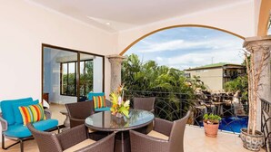 A balcony with wicker furniture, a glass table, and a view of a waterfall and greenery.