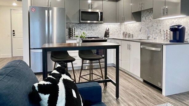 Een moderne keuken met een ingebouwde magnetron, een roestvrijstalen koelkast en een vaatwasser. Er staat een houten eettafel met twee zwarte barkrukken.