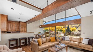 A spacious living room with a large window, a wooden ceiling beam, and a kitchen area with wooden cabinets and bar stools. - Landmark 702 - 3 Bedroom Diamond (Vail)