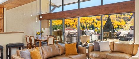 A spacious living room with leather sofas, a wooden coffee table, and large windows offering a view of autumn foliage.