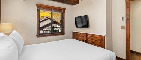 Ein Schlafzimmer mit einem Bett, einer Holzkommode, einem an der Wand befestigten Fernseher und einem Fenster mit Blick auf ein Gebäude.