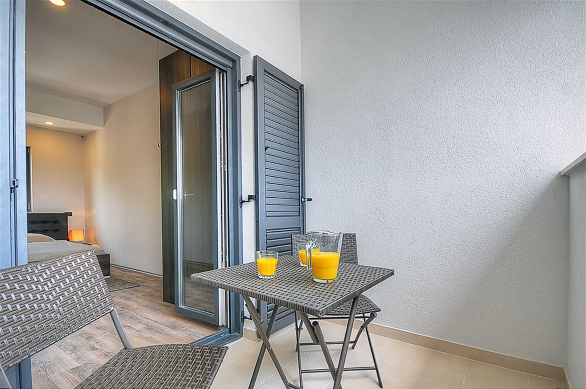 A small balcony with a table and chairs, two open sliding doors leading to a bedroom, and a bed with a headboard visible in the background.
