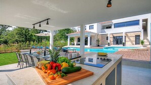 An outdoor kitchen with a wooden cutting board, various vegetables, and a built-in grill. In the background, there is a swimming pool and a modern house with a stone facade. - Villa Dahlia Belle with Pool (Višnjan)