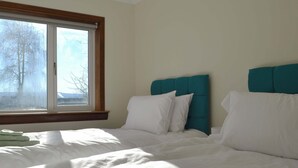 Une chambre avec une grande fenêtre, un lit avec du linge de lit blanc, une tête de lit turquoise et une vue sur les arbres à l’extérieur.