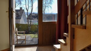 Une pièce avec un escalier en bois, une porte-fenêtre coulissante donnant sur un patio équipé d’une chaise, et une vue sur les arbres et les maisons à l’extérieur.
