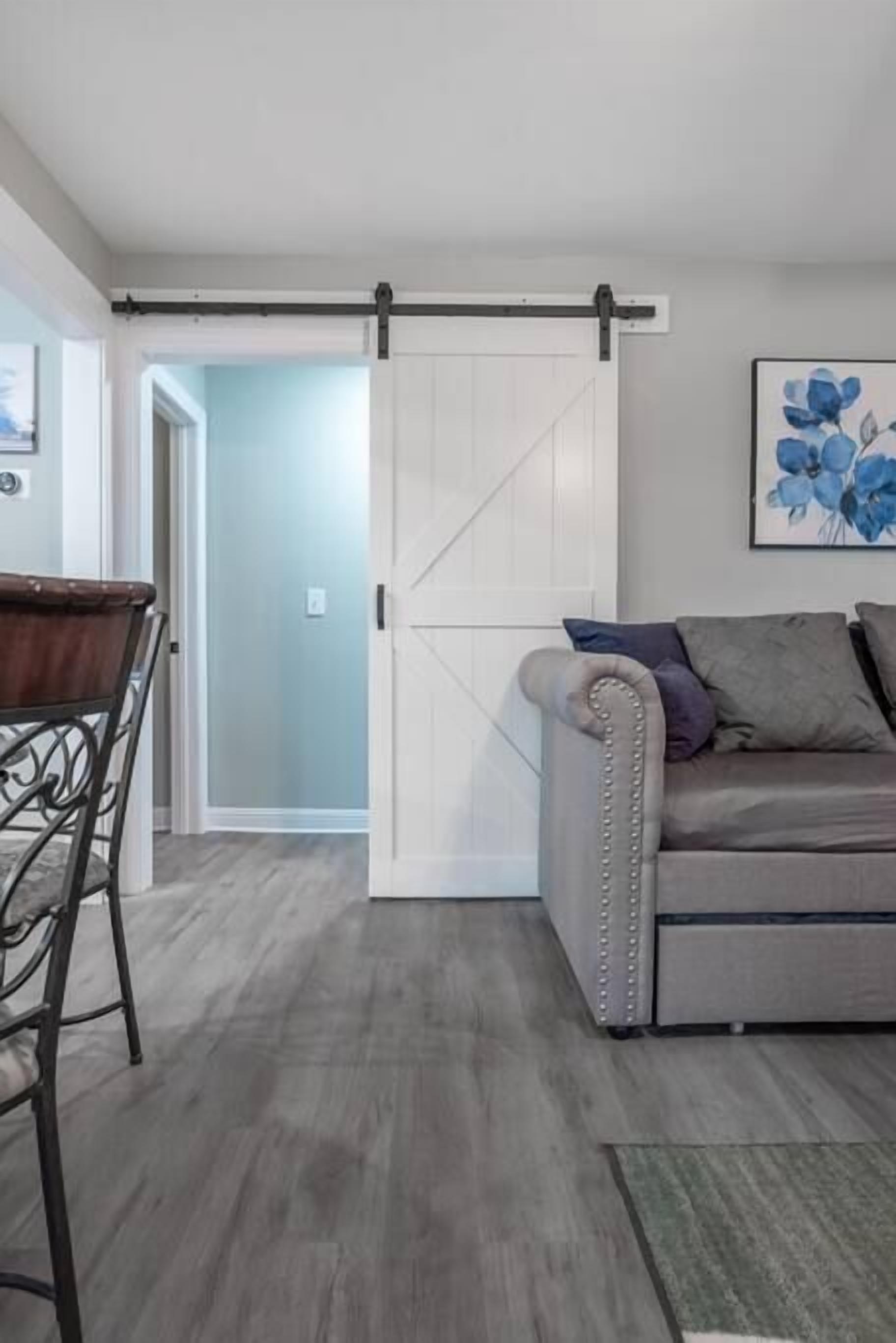 Un salon moderne avec une porte coulissante de style rustique, un canapé gris agrémenté de coussins, une table à manger en bois avec des chaises en métal, et une œuvre d’art florale encadrée accrochée au mur.