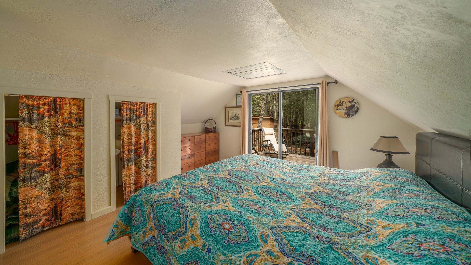 A bedroom with a large bed, a dresser, a chair, and a balcony with a view of trees.